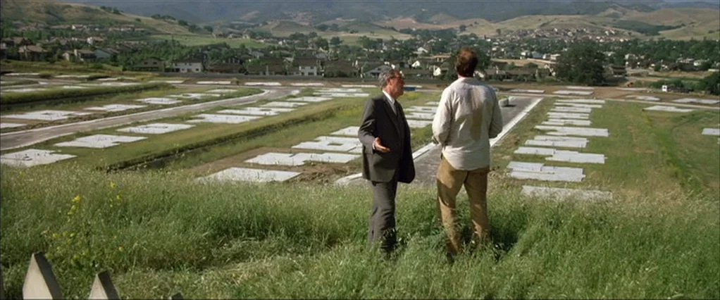 Poltergeist movie still: wide — A grassy slope overlooks a vast suburban development site with numerous white concrete foundations. Two men in…
