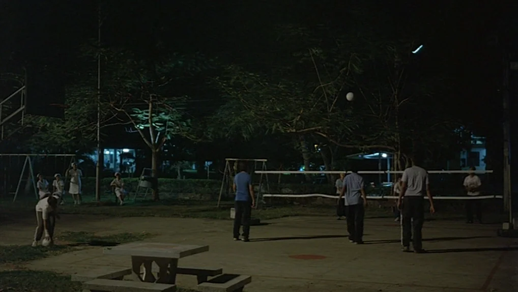 Syndromes and a Century (2006) movie still: wide — A nighttime outdoor paved area features a volleyball net and nearby trees. Several individuals…