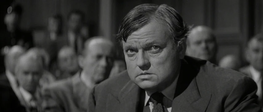 Compulsion (1959) movie still: closeup — A crowded public gallery fills a dimly lit space with wood paneled walls. A middle-aged man in a dark suit…