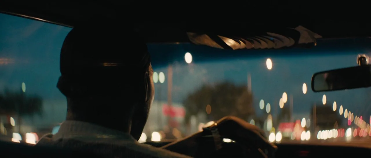 Moonlight (2016) movie still: medium — The interior of a car at night looking out onto a city street. A man sits behind the steering wheel watching…