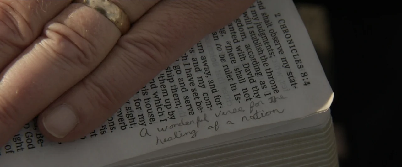 Reagan (2024) movie still: closeup — A close-up view shows a person's hand resting on the page of an open bible. A handwritten note in pencil at the…