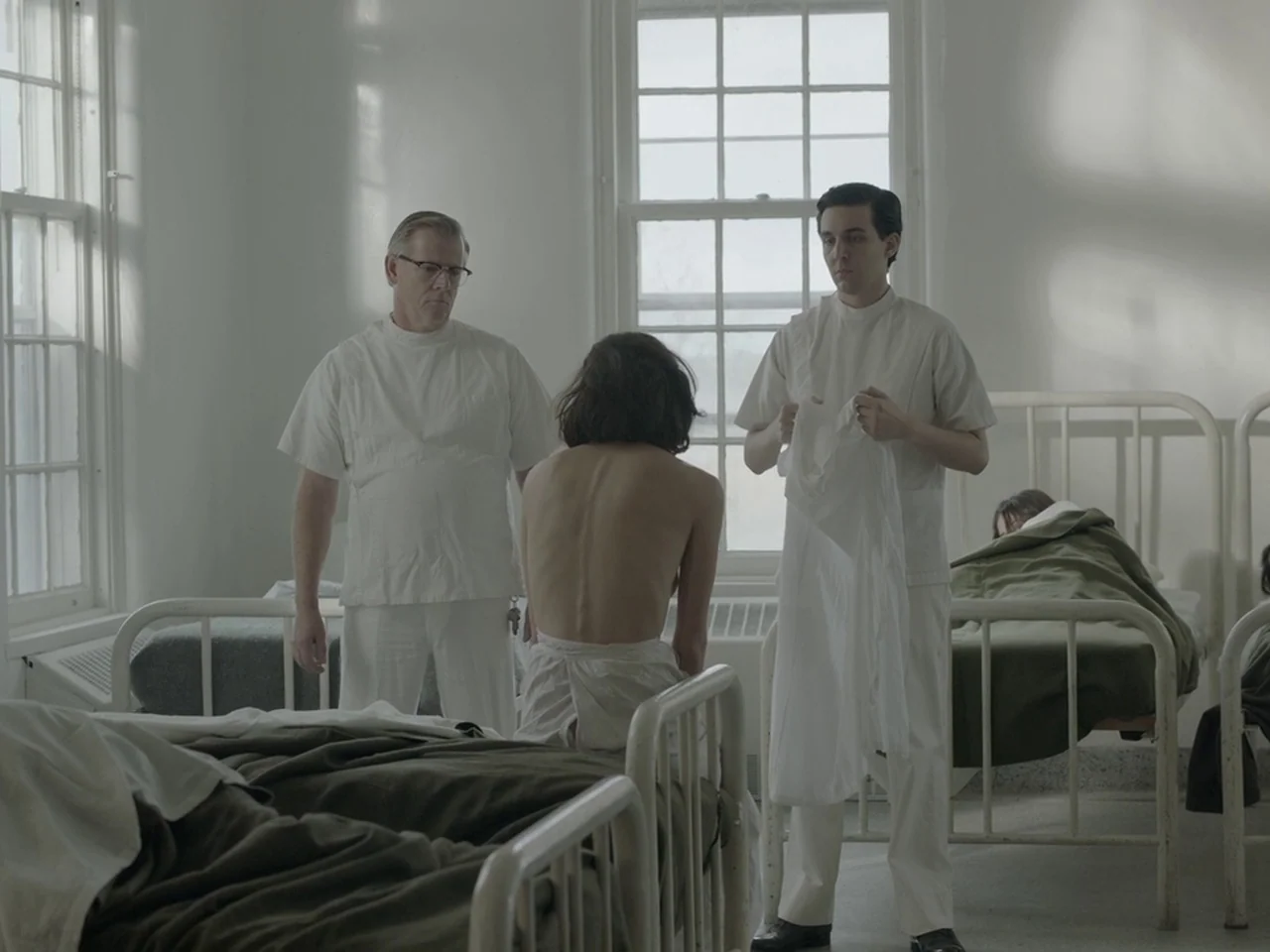 The Mountain (2018) movie still: medium — A sterile hospital ward features high ceilings, white walls, and rows of metal beds. Two men in white…