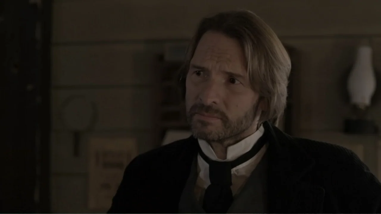 Dawn Rider (2012) movie still: medium — A 1900s indoor setting with wooden paneled walls and a blurred lamp in the background. A man with…