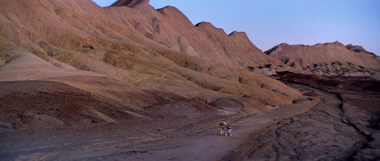 Return Of The Jedi (1983) movie still: wide — A vast, arid desert landscape features towering, eroded rock formations and a dirt track. A golden…