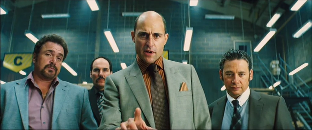 Kick Ass (2010) movie still: medium — A utilitarian industrial space with concrete block walls and hanging fluorescent light fixtures. Four men in…