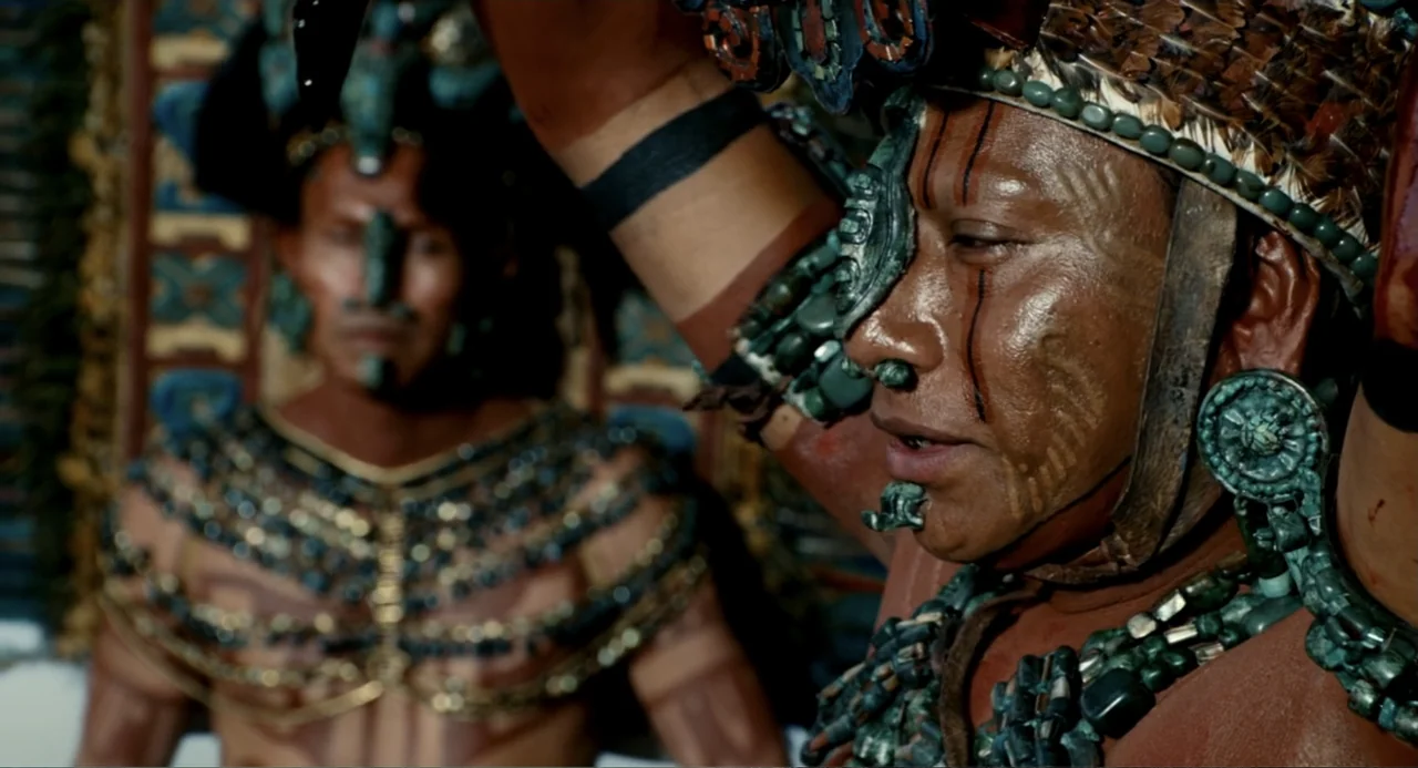 Apocalypto (2006) movie still: medium — An outdoor plaza features two men decorated in traditional ancient indigenous ceremonial regalia. The…