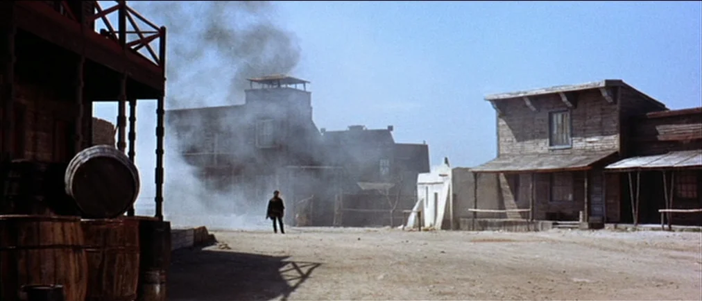 Fistful Of Dollars (1964) movie still: wide — An arid, unpaved street runs through a collection of weathered, wooden frontier buildings. Thick grey…