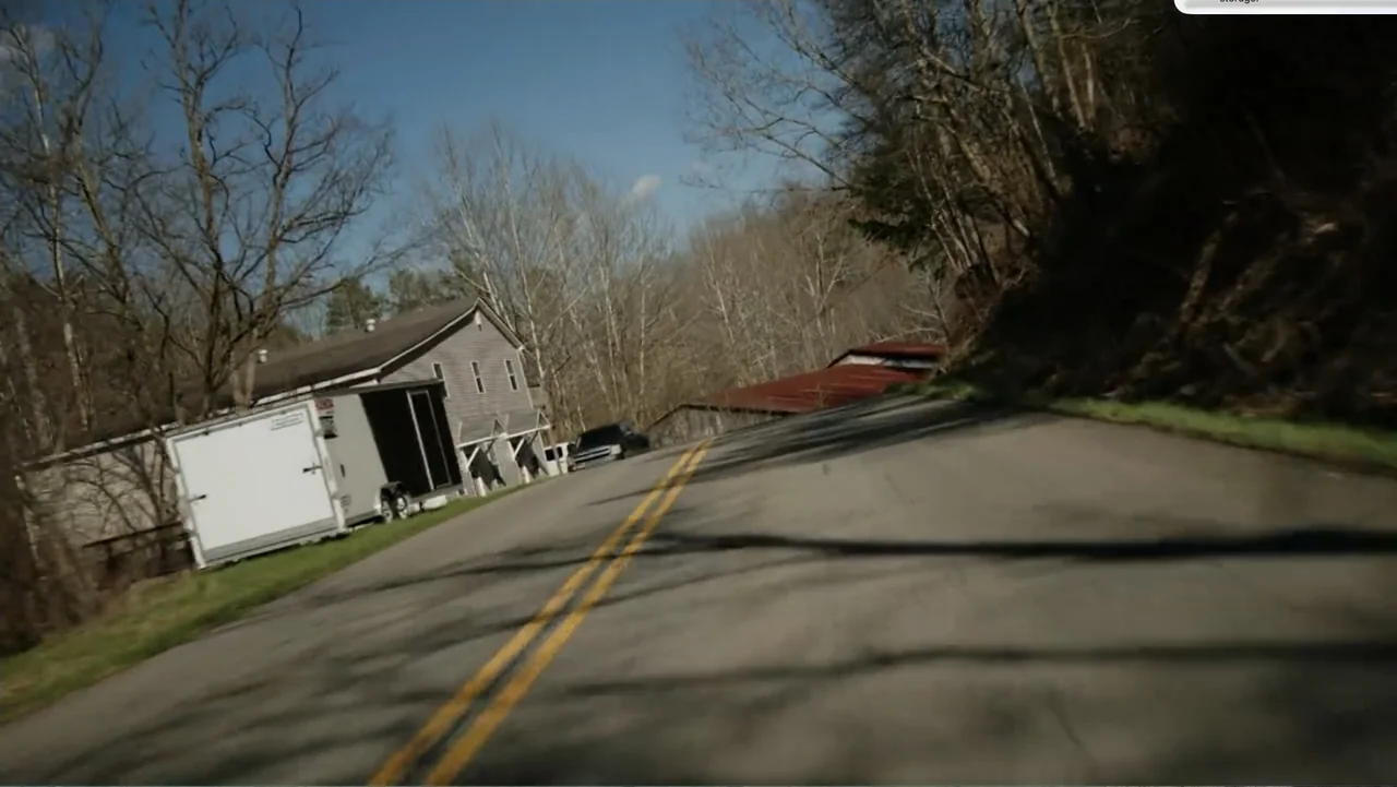 Murder at Carter County Mines (2023) movie still: wide — A rural asphalt road passes by a grey building and a white utility trailer. Bare trees line…
