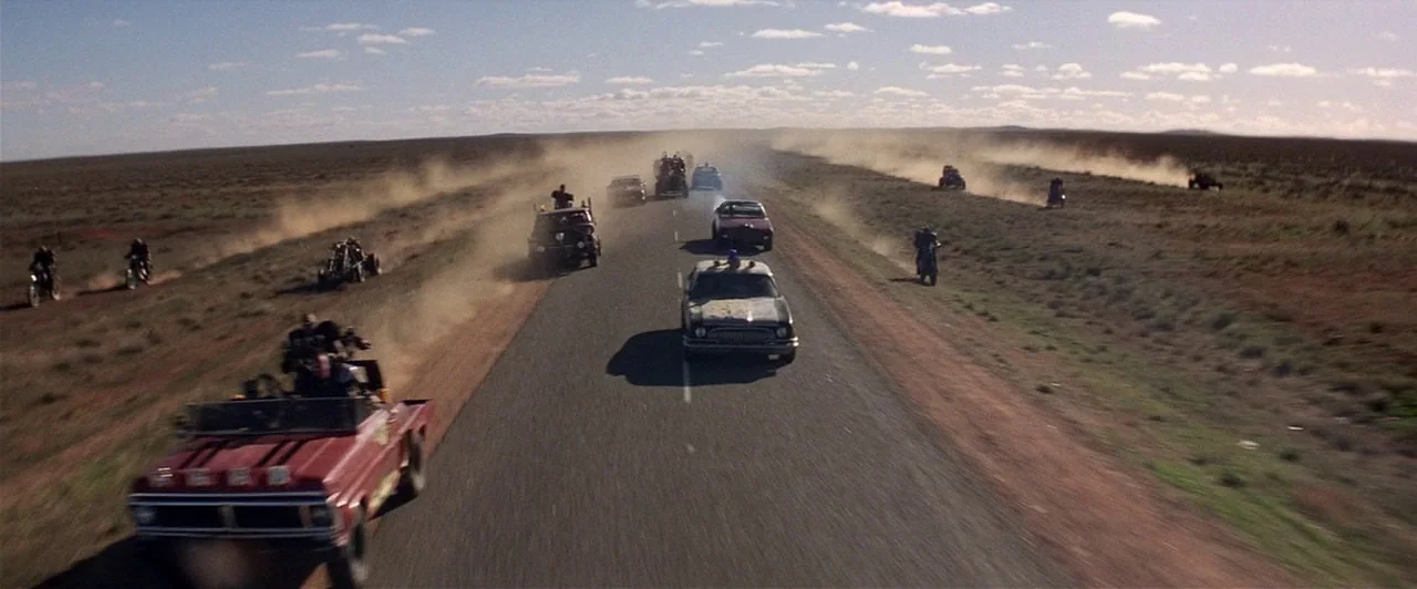 Mad Max 2 (1981) movie still: wide — A long, flat stretch of paved road cuts through an expansive, dry, arid landscape. Several modified vehicles,…