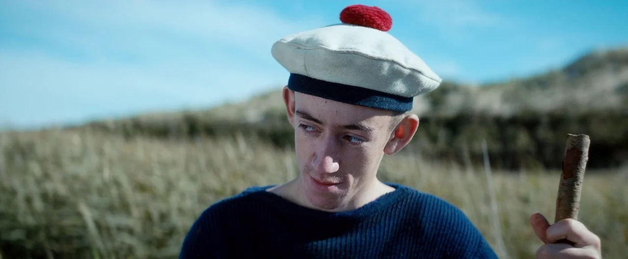 Slack Bay (2016) movie still: medium — A coastal dune area with tall, dry grass stretching toward a distant sandy hill. A young man wearing a…