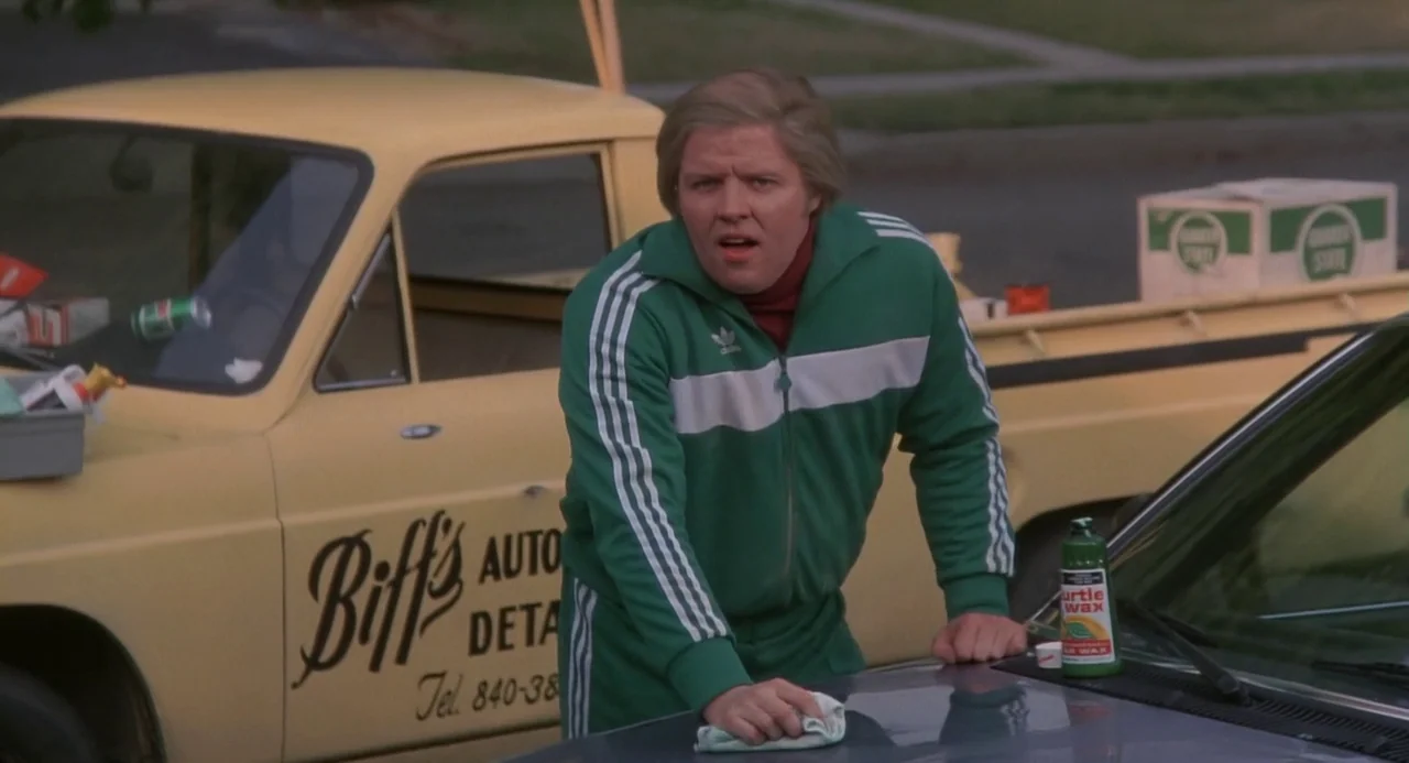 Back to the Future (1985) movie still: medium — A suburban residential setting with a yellow pickup truck parked near a clean car. A man in a green…