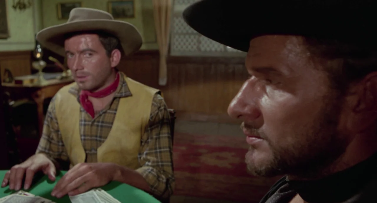 Johnny Colt (1966) movie still: medium — A dimly lit room with wood-paneled walls and a green card table. Two men in cowboy attire sit opposite each…