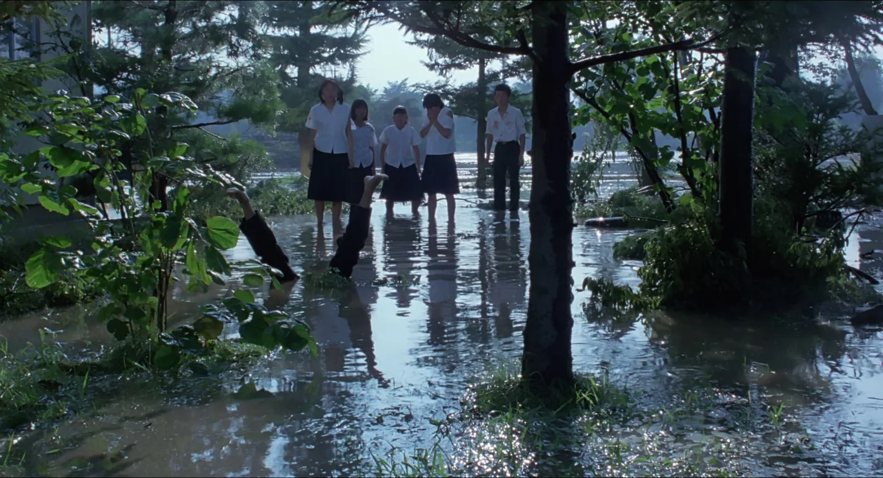 Typhoon Club (1985) movie still: wide — Five teenagers stand in a shallow flooded wooded area among submerged trees and debris. A pair of legs in…