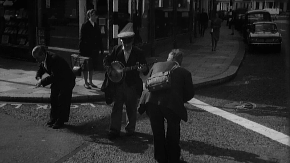 Repulsion (1965) movie still: fullbody — An urban intersection with a stone-paved corner and storefronts in the background. Three men stand on the…