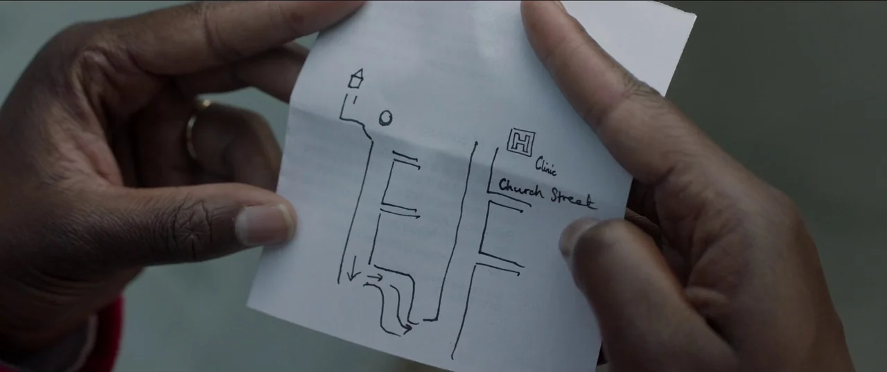 His House (2020) movie still: closeup — A pair of dark-skinned hands hold a small white scrap of paper. A simple hand-drawn map with street labels…