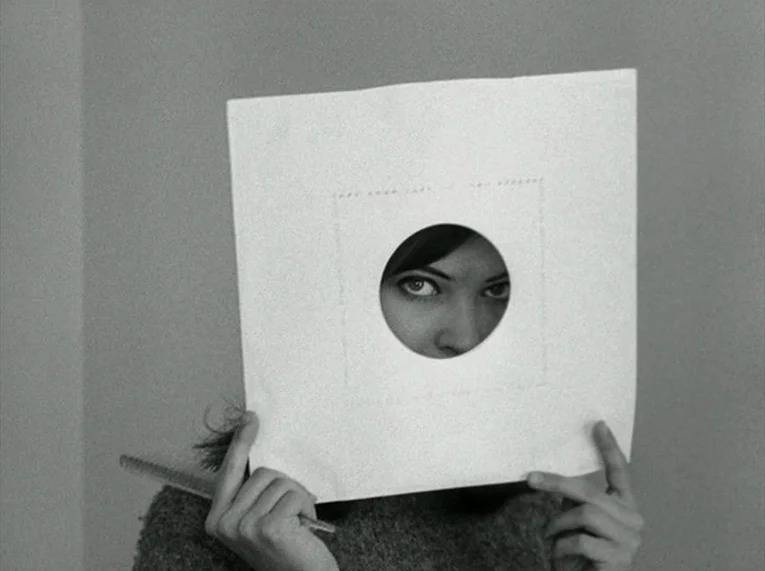 Le Petit Soldat (1963) movie still: closeup — A simple interior space with a plain, neutral-toned wall in the background. A young woman holds a…