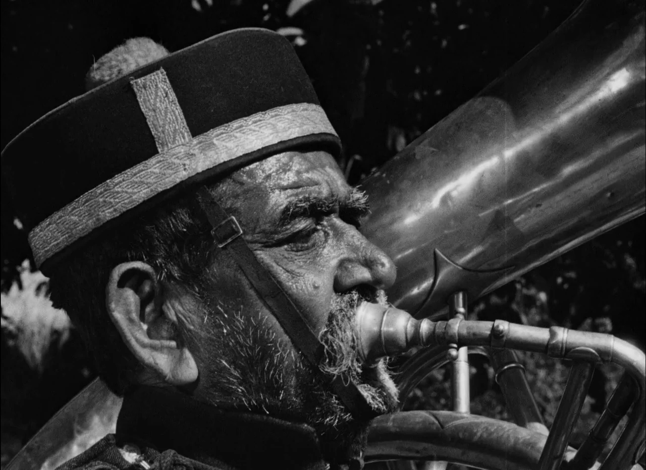 Pather Panchali (1955) movie still: closeup — An elderly man plays a large brass horn outdoors in natural sunlight. He wears a peaked uniform cap…