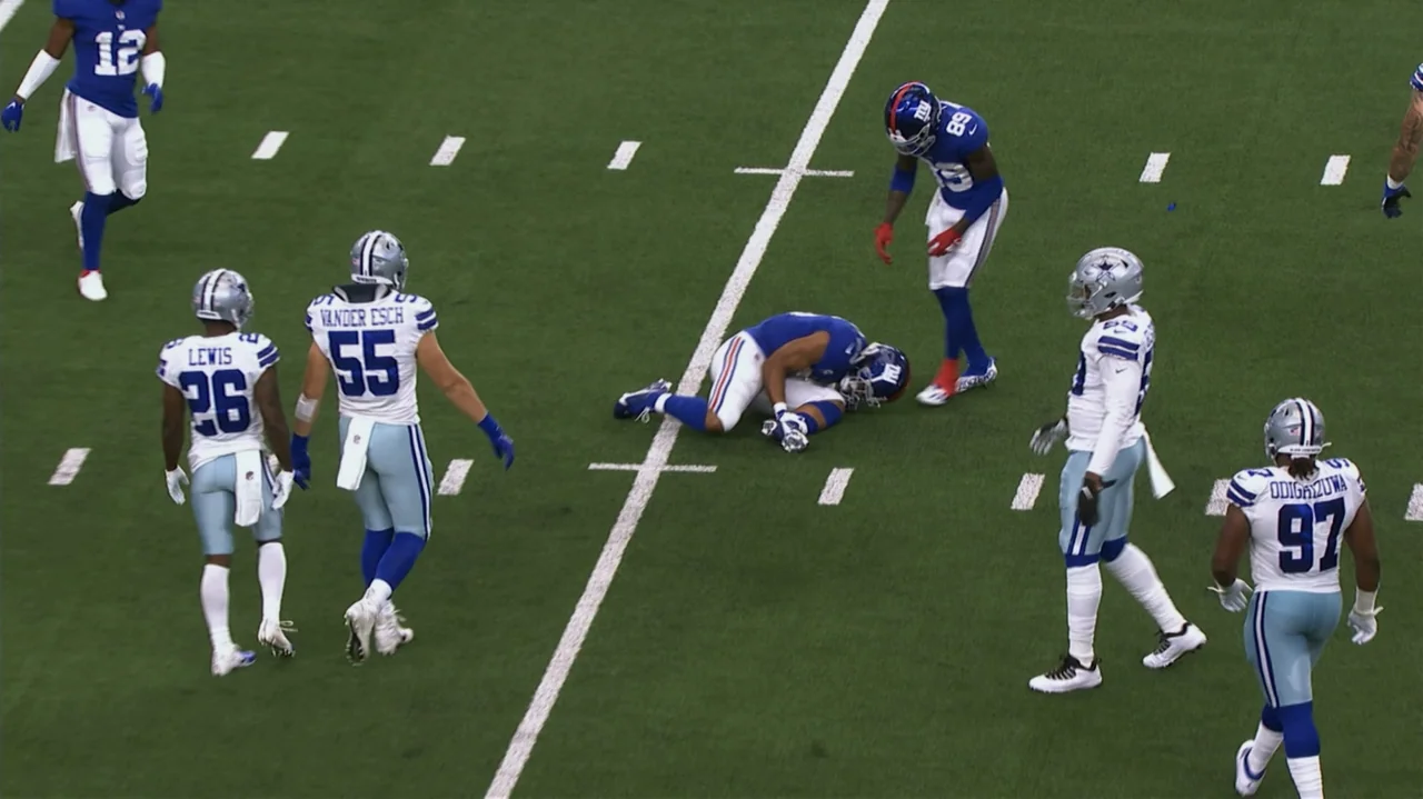 SAQUON (2025) movie still: wide — A football field covered in green turf has white painted yard lines visible. One football player lies prone on the…