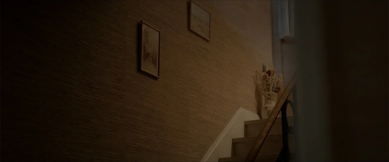 An Endless Wedding (2025) movie still: wide — A residential staircase landing features textured beige wallpaper and two small framed pictures. A…