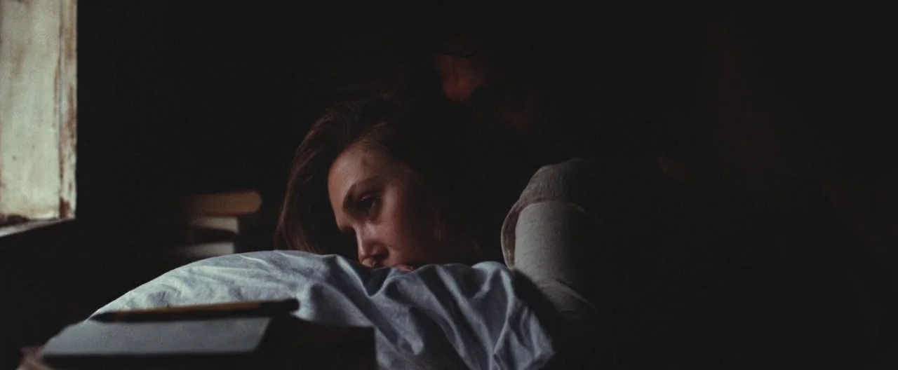 Martha Marcy May Marlene (2011) movie still: medium — A dimly lit room features a bed positioned near a window with natural light. A woman lies on a…