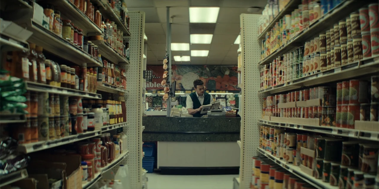 The Monkey (2025) movie still: wide — An aisle in a grocery store is lined with shelves containing many canned goods. A young man wearing a green…