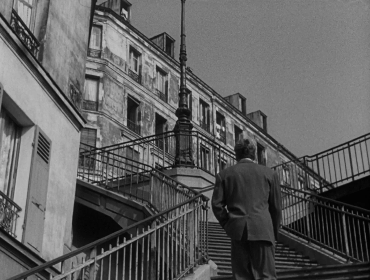 Orpheus (1950) movie still: fullbody — A weathered multi-story urban residential building rises above a large concrete staircase. A man in a suit…