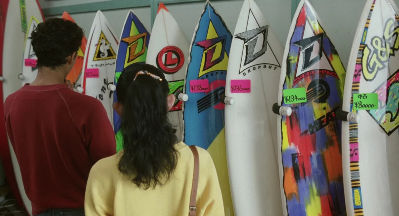 A Scene at the Sea (1991) movie still: medium — A surf shop interior features a row of colorful surfboards displayed on a wall. A man and a woman…