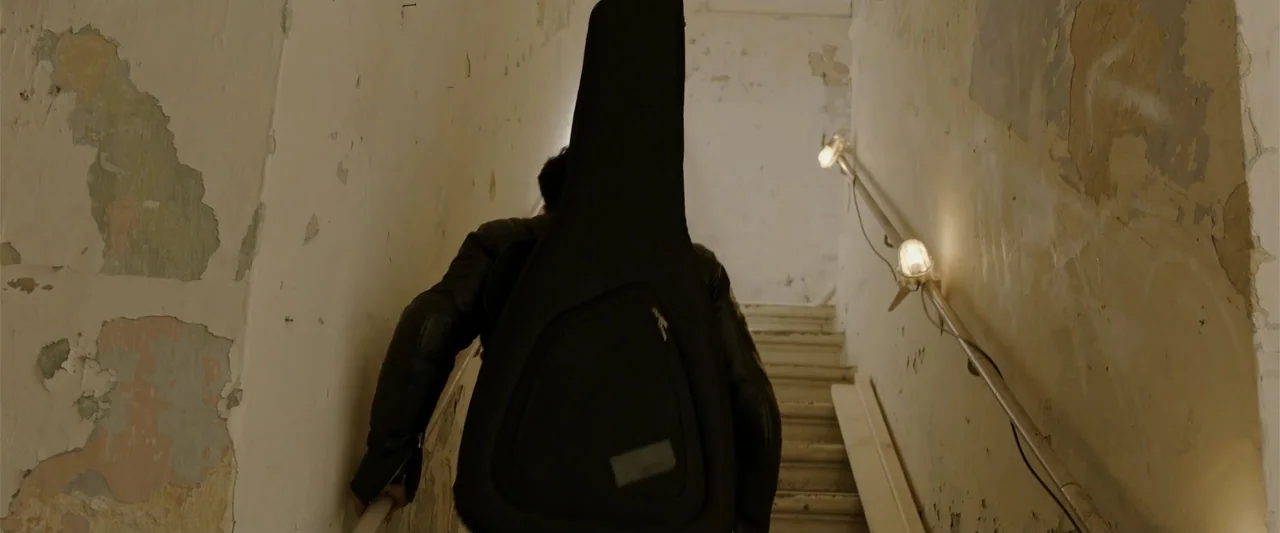 Hounds of War (2024) movie still: medium — A narrow residential stairwell features walls with peeling paint and exposed plaster patches. A person…