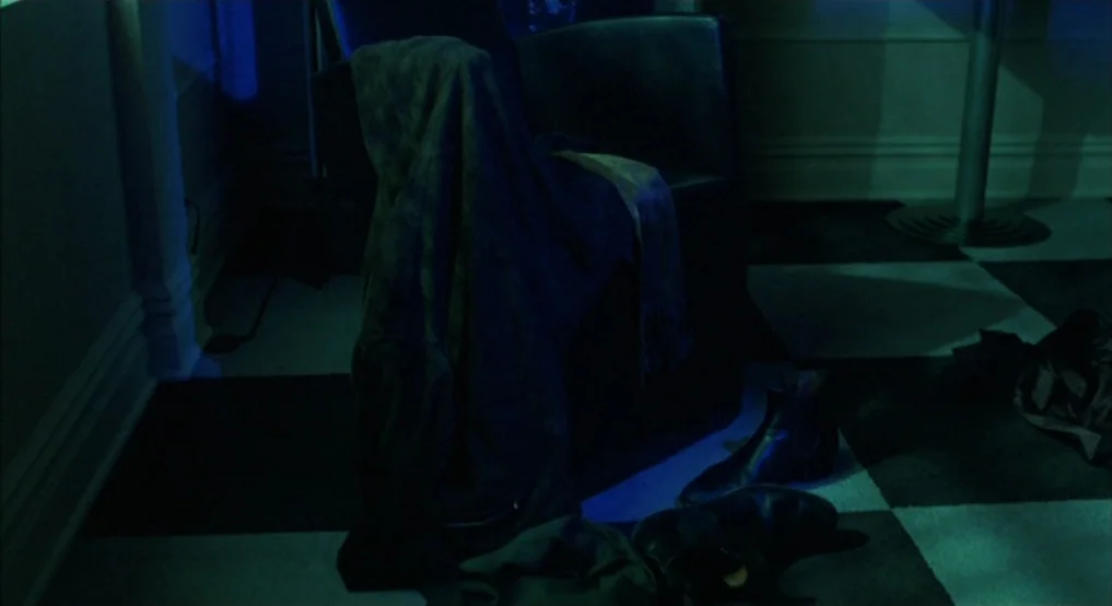 Light Sleeper (1992) movie still: medium — A dark room features a black checkered tile floor and a single upholstered armchair. A coat is draped…