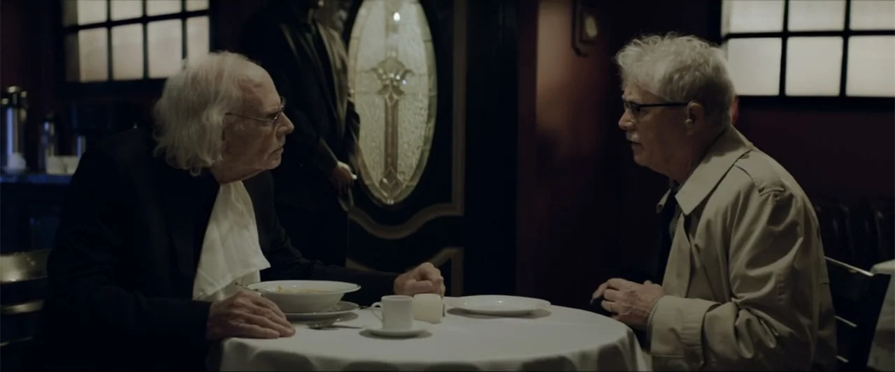 Gender Under Attack (2018) movie still: medium — A dimly lit restaurant booth features dark walls and a round table with white linens. Two elderly…