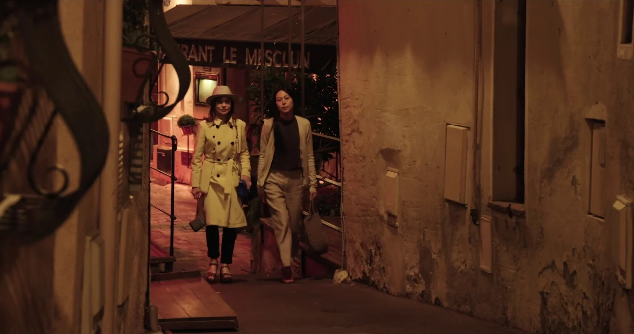 Claire’s Camera (2017) movie still: fullbody — A narrow stone-walled passage leads toward the illuminated entrance of a restaurant at night. Two…
