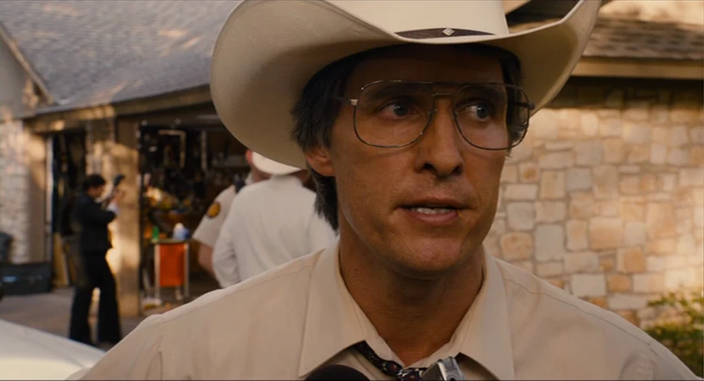 Bernie (2011) movie still: closeup — A residential exterior driveway features a stone house and a partially open garage door. A man in a cowboy hat…