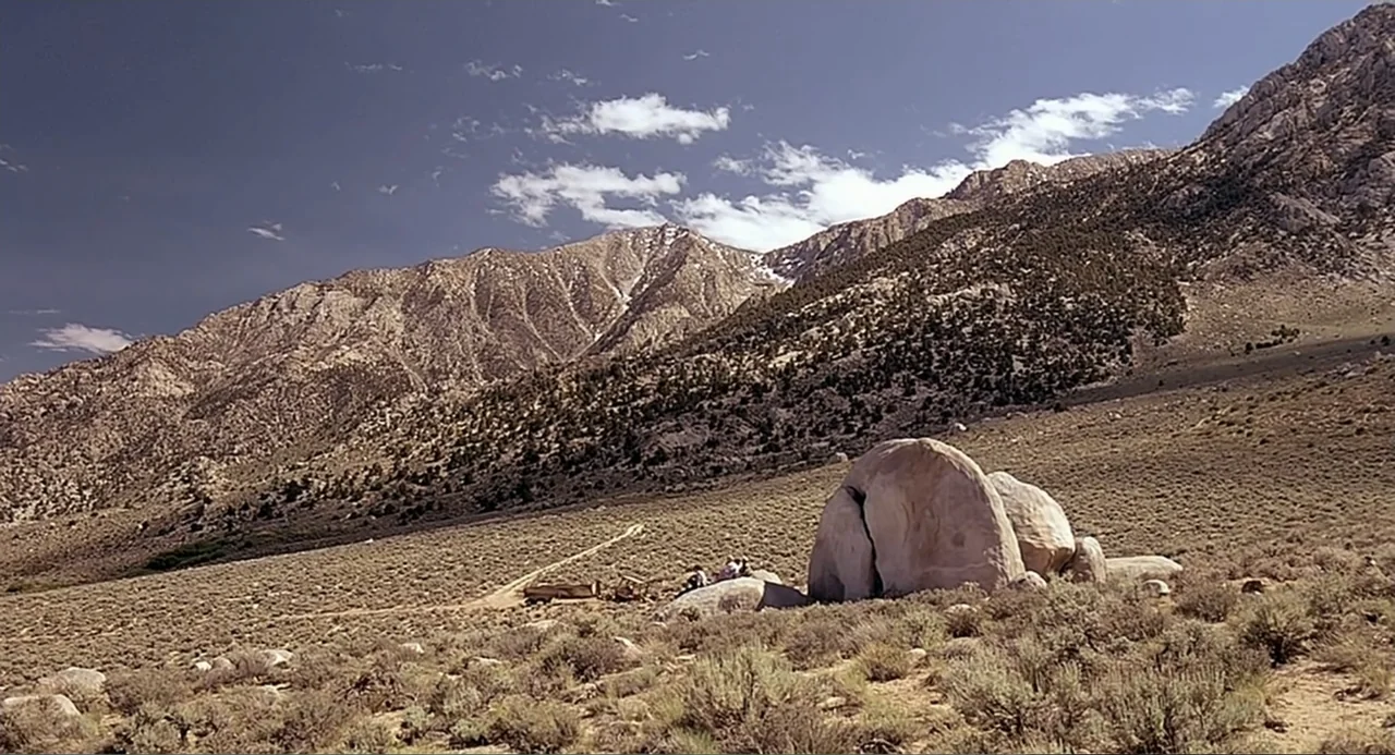 Tremors (1990) movie still: wide — A vast arid landscape features scattered shrubs and a large light-colored boulder. Distant mountains rise against…