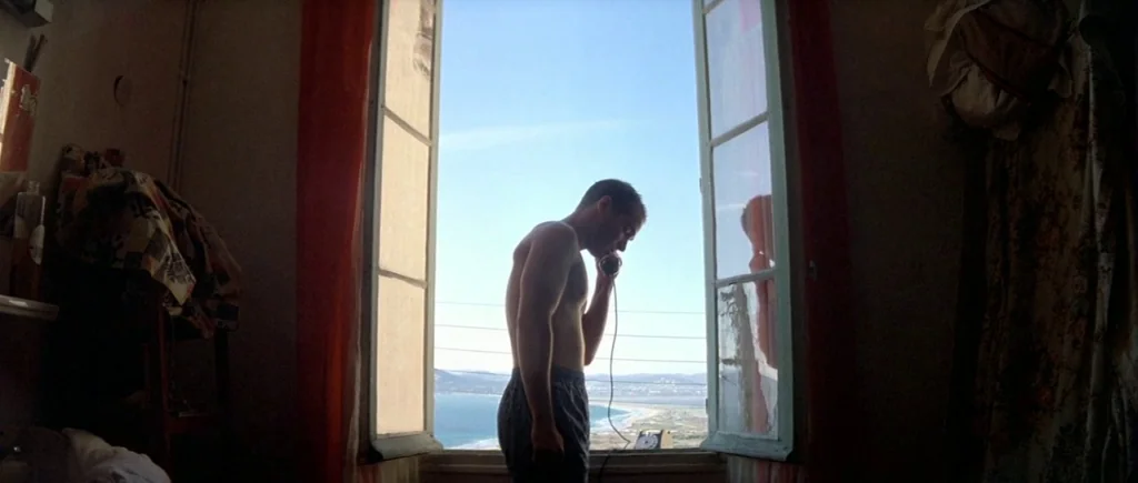 The Big Blue (1988) movie still: fullbody — A sparsely furnished room with a large open window overlooking a coastal horizon. A shirtless man stands…