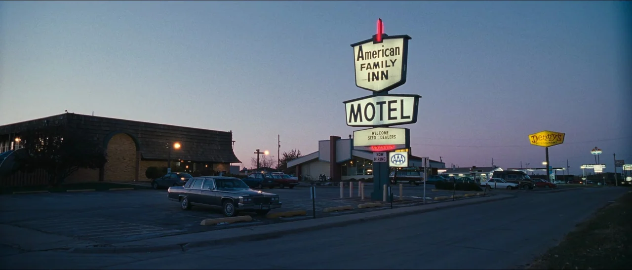 Election (1999) movie still: establishing — A paved motel parking lot sits under a clear blue twilight sky at dusk. A tall illuminated sign towers…