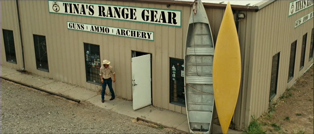 No Country For Old Men movie still: wide — A single-story building with corrugated tan metal siding has a sign for range gear. A man in a cowboy hat…