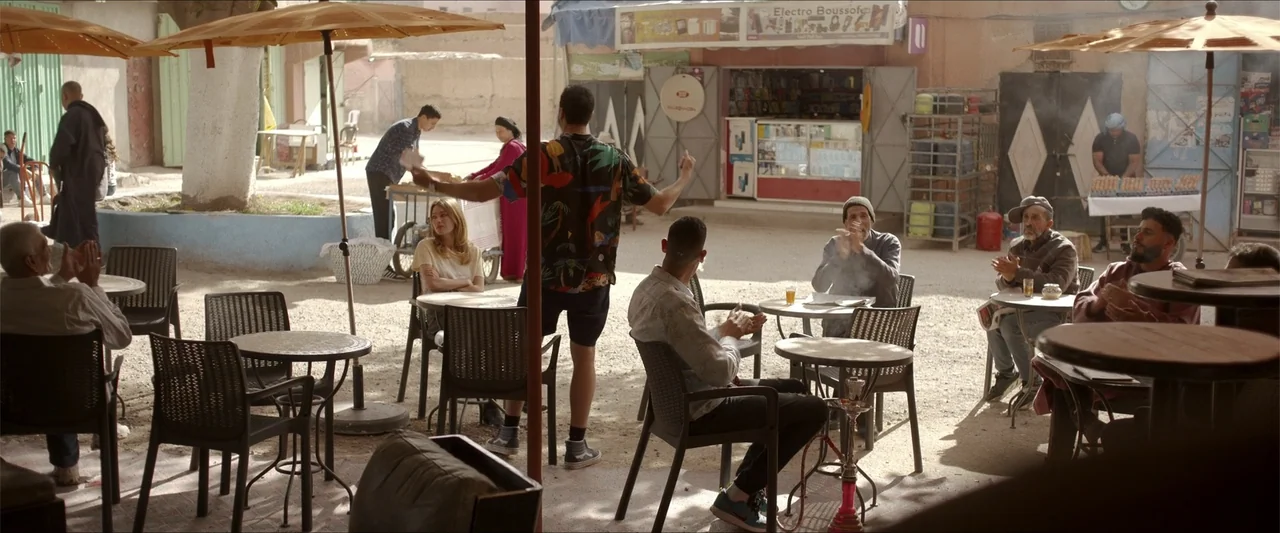 An Endless Wedding (2025) movie still: fullbody — A dusty outdoor plaza filled with several small cafe tables and plastic chairs. A group of people…