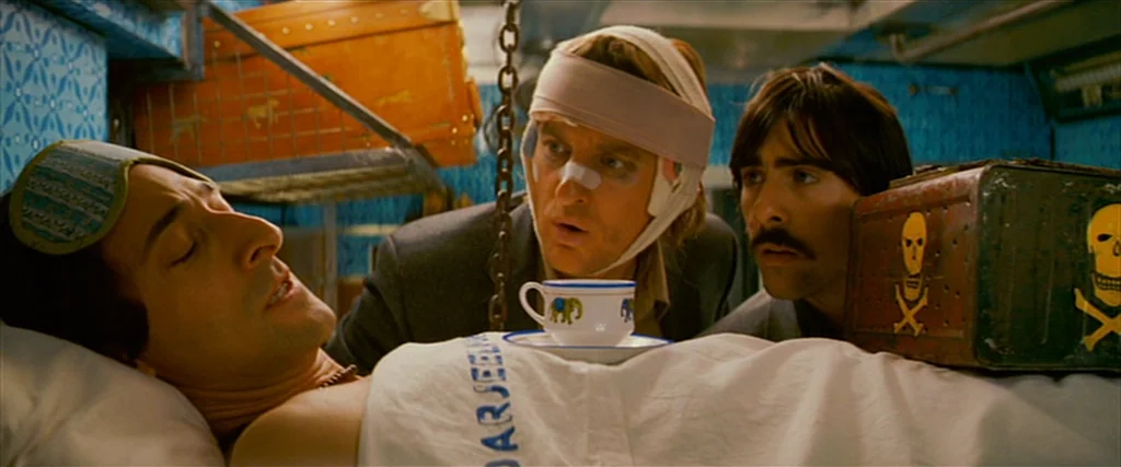 The Darjeeling Limited (2007) movie still: medium — A cramped train compartment with bright blue patterned walls and leather luggage overhead. Three…