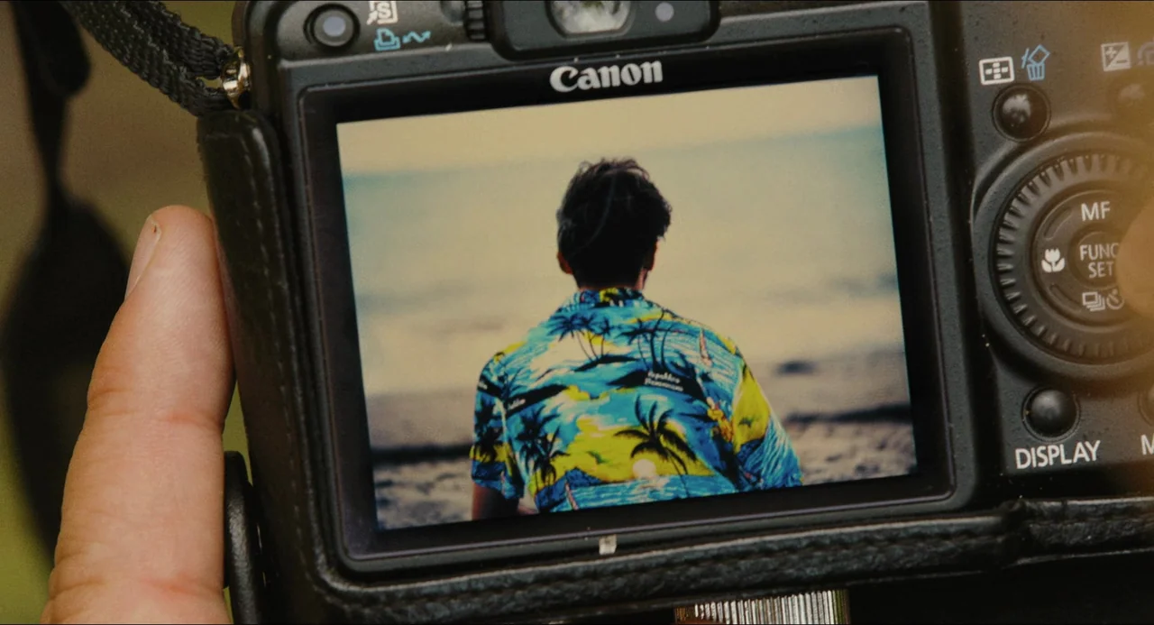 Afire (2023) movie still: closeup — A close-up view shows a Canon digital camera screen displaying a photo of a man. The man stands with his back to…