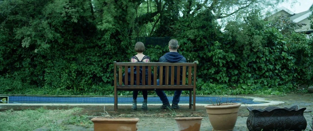 The Empty Man (2020) movie still: fullbody — A backyard contains a swimming pool bordered by thick, dense green ivy walls. A man and woman sit side…