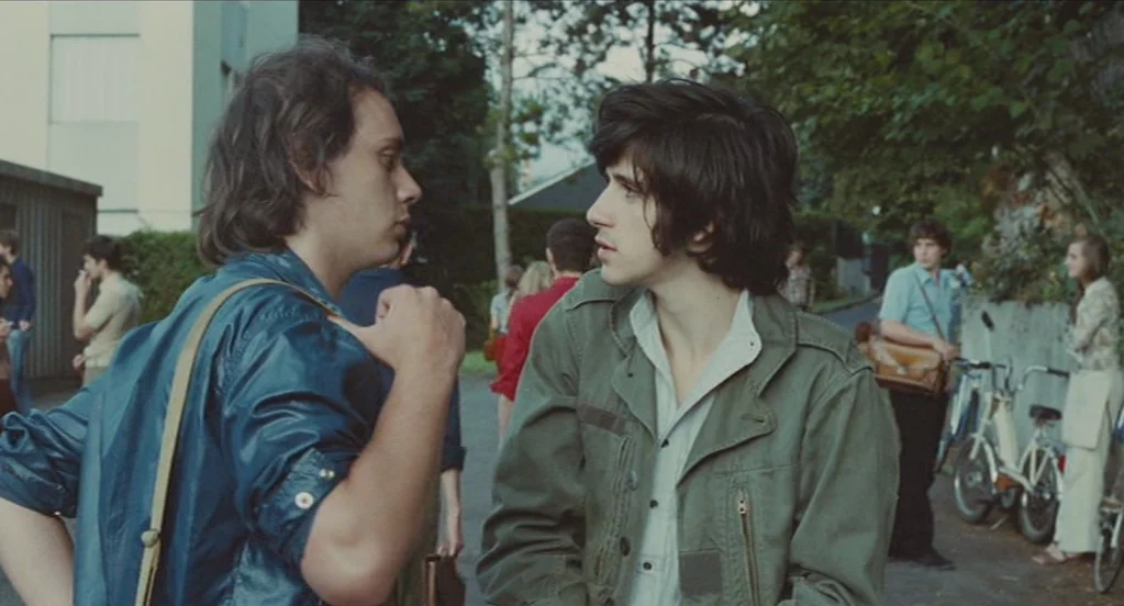 Something in the Air (2012) movie still: medium — Two young men converse in a paved lot near apartment buildings and foliage. Other people and a…