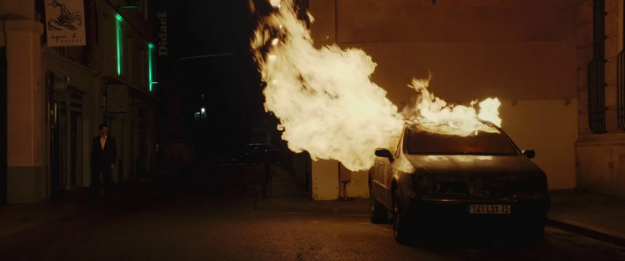 Nocturama (2016) movie still: wide — A dark, empty city street at night features a car engulfed in bright fire. A solitary man in a suit stands on…