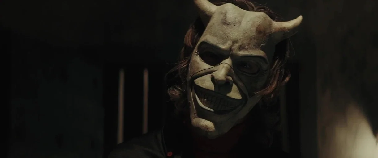 The Black Phone (2021) movie still: closeup — A dimly lit room with blurred vertical bars in the dark background. A man wearing a devil mask stares…