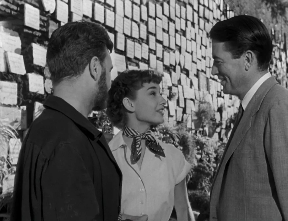 Roman Holiday (1953) movie still: medium — A stone wall is covered in numerous small white memorial plaques in orderly rows. Three people stand in a…