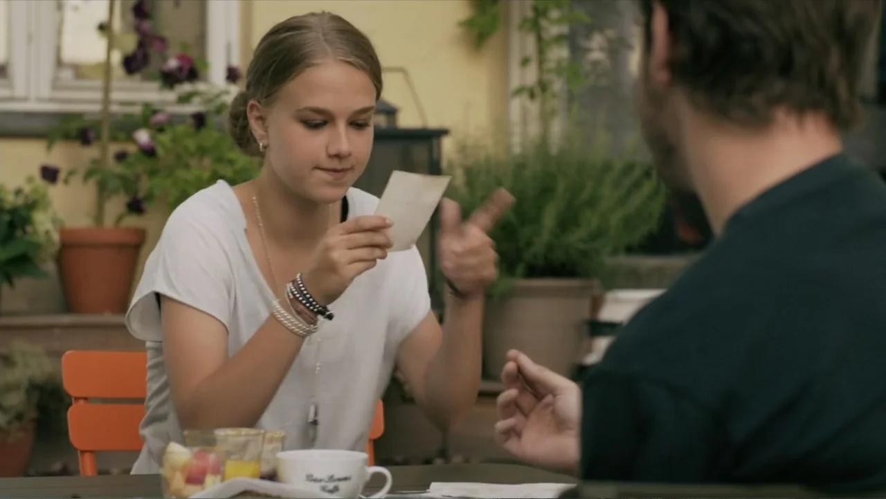 Copenhagen (2002) movie still: medium — A sunny outdoor patio with potted plants and table settings in the background. A young woman holds up a…