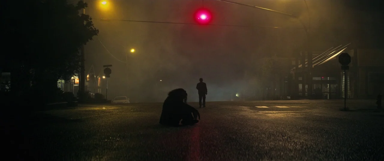 Come To Daddy (2019) movie still: wide — A wet intersection at night is thick with swirling fog and yellow street lighting. One person sits in the…