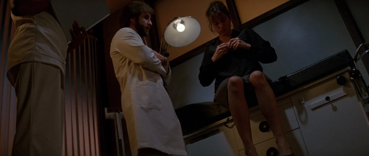 The Entity (1982) movie still: medium — A dimly lit medical room features a tall metal examination lamp and cabinet storage. A doctor in a lab coat…