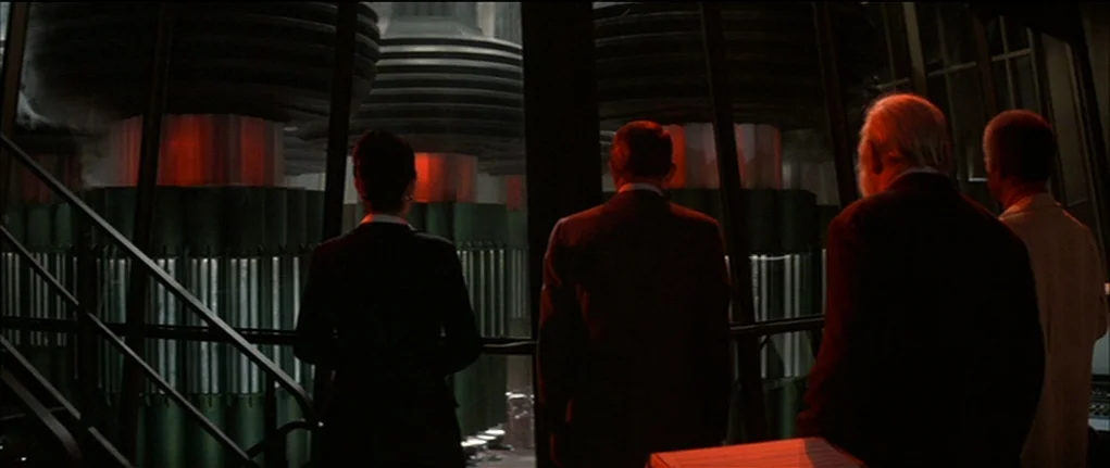 Demon Seed (1977) movie still: wide — A dark observation deck overlooks large, segmented industrial machinery with glowing red internal cores. Four…