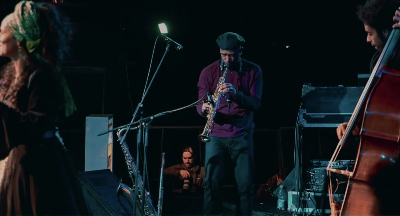 Cairo Jazzman (2017) movie still: medium — A dimly lit musical performance space featuring three musicians on a dark stage. A saxophonist stands…