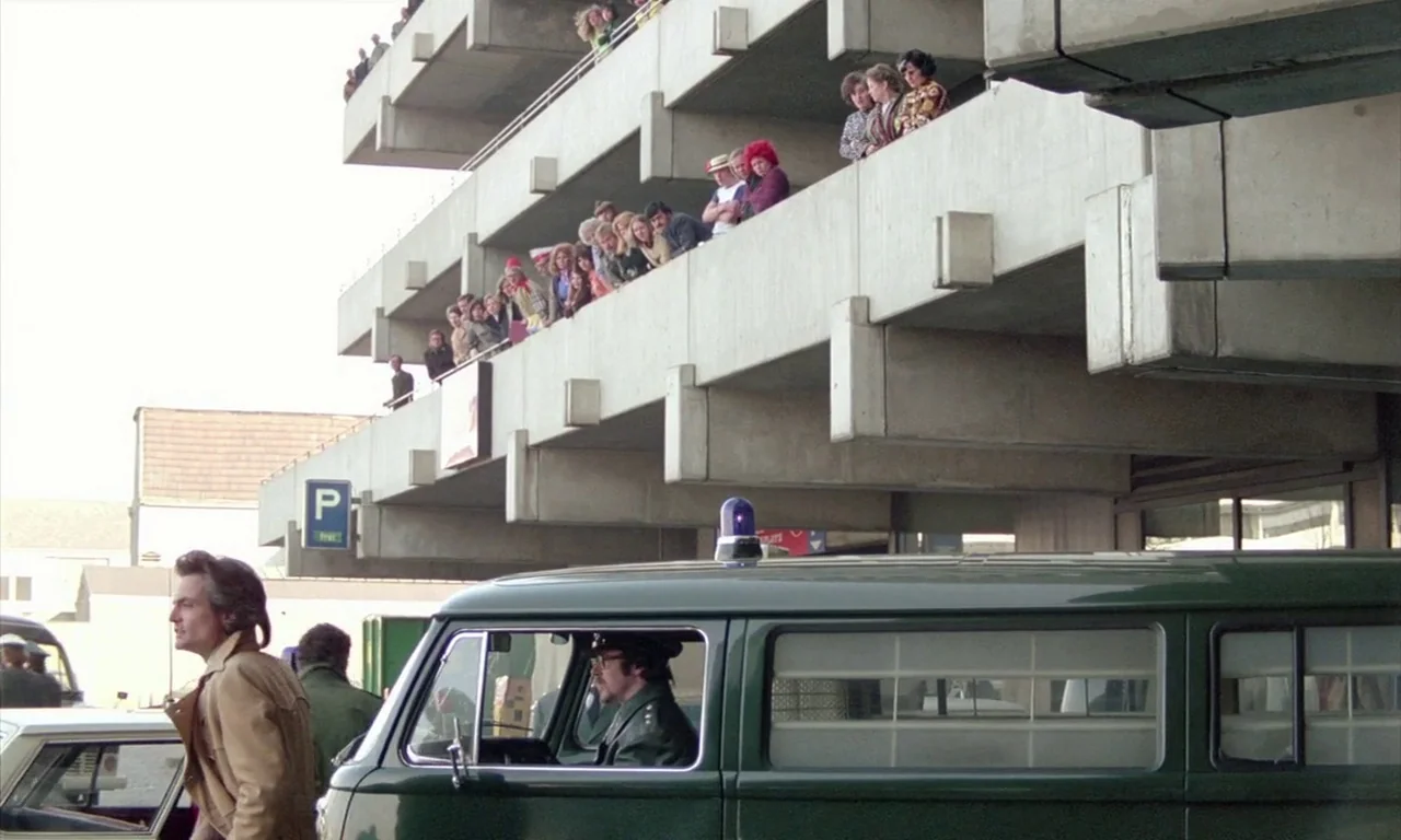 The Lost Honour of Katharina Blum (1975) movie still: wide — A modern concrete parking garage structure features a crowd of people watching from…